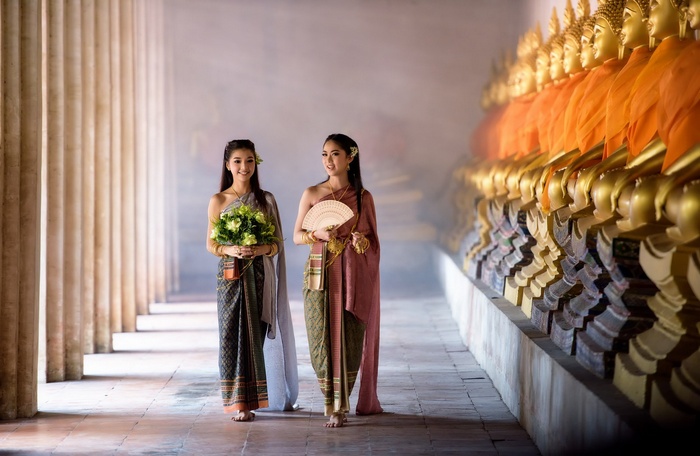 Two women in traditional Thai clothing walking near Buddha statues