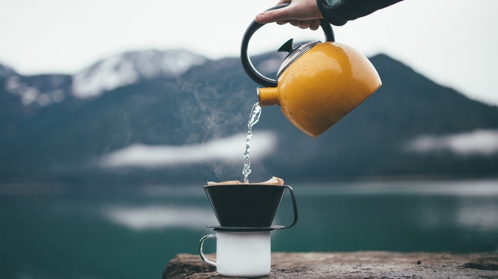 Pouring hot water from a kettle into a coffee filter over a cup with mountains and a lake in the background.
