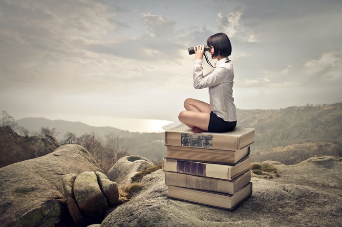 Woman with binoculars sitting on books on a rocky hill