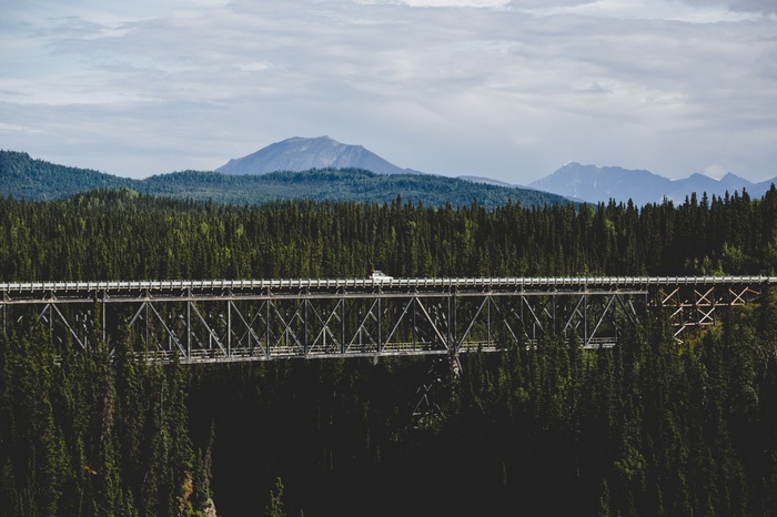 A white car drives across a bridge over a forest with mountains in the background.