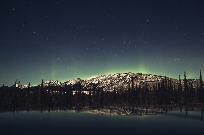 Aurora Borealis over snow-capped mountains and forest reflected in water