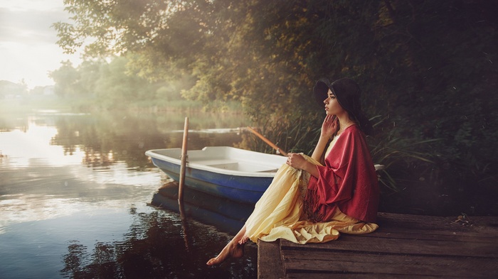 Woman in hat and dress sitting by a boat on a river