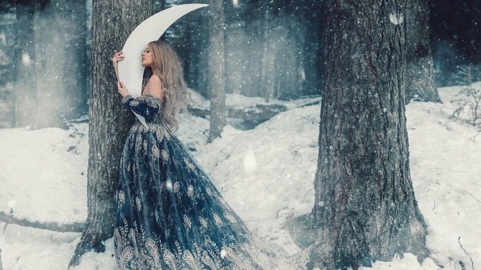 Woman in a blue dress holding a crescent moon in a snowy forest