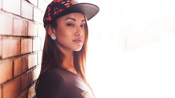 Asian woman wearing a hat leaning against a brick wall