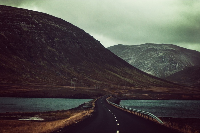Road through mountains beside a lake under stormy clouds
