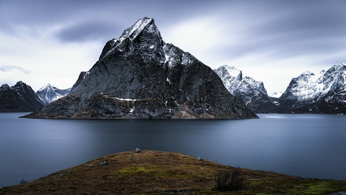 Snow-capped mountains rise from a calm lake under a cloudy sky