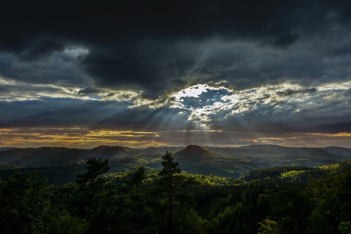 Sunlight beams through dark clouds over a forested mountain landscape