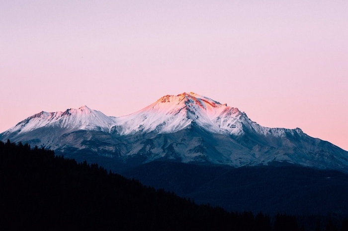 Snow-capped mountains at sunset with a dark forest foreground