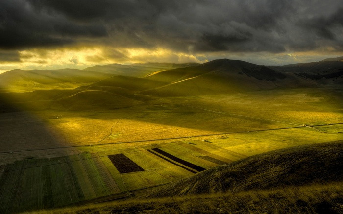 Sunlight beams through dark clouds over rolling hills and fields