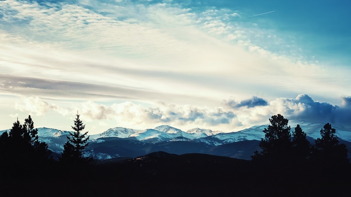 Silhouette of trees against snowy mountains and clouds