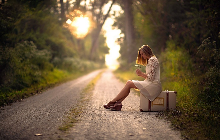 Woman sitting on suitcase on a road with flowers