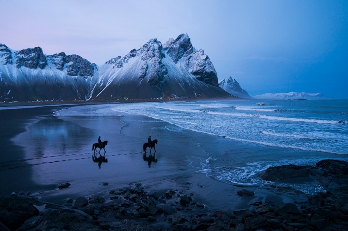 Two people on horses on a beach with mountains