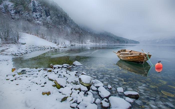 Wooden boat moored on a misty winter lake with snow-covered mountains