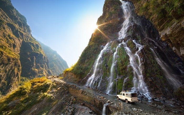 Waterfall cascading down a mountain with a vehicle on a dirt road in Nepal