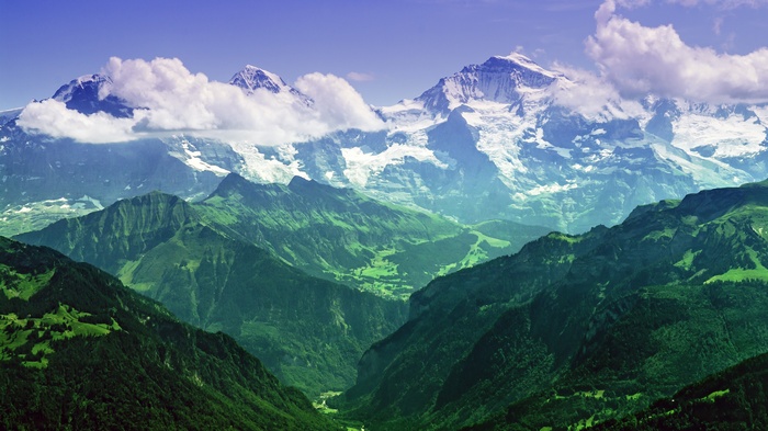 Snow-capped mountains rise above green valleys under a blue sky.