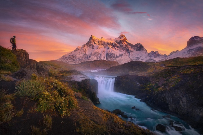Hiker overlooking a waterfall and snowy mountains at sunset