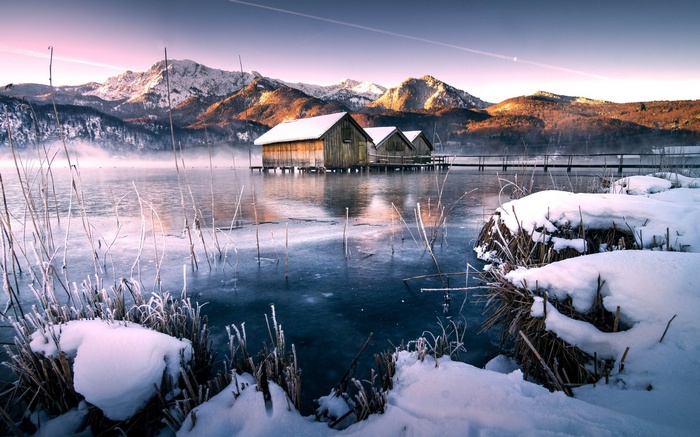 Wooden boathouses on a frozen lake with snow-capped mountains