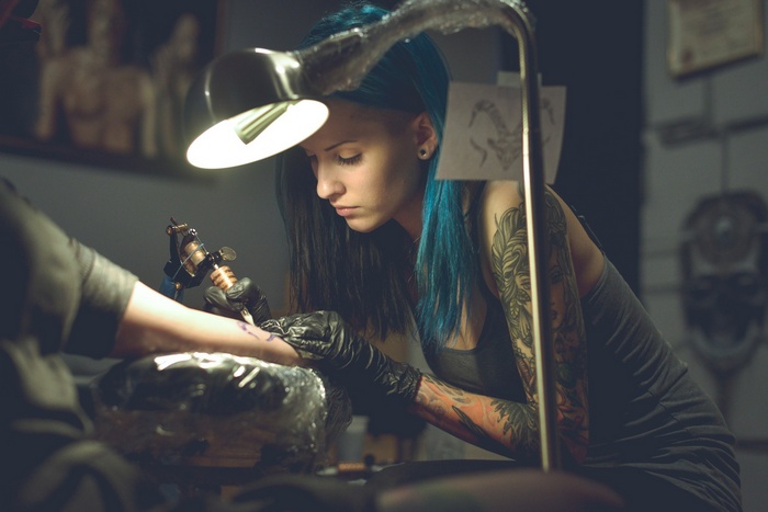 Woman with blue hair tattooing an arm