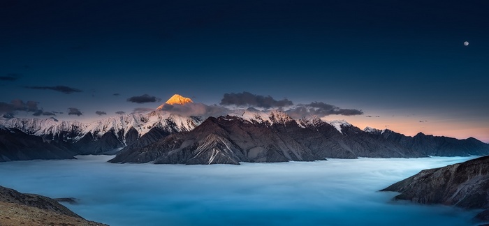 Mountains covered in snow and mist under a blue sky with the moon