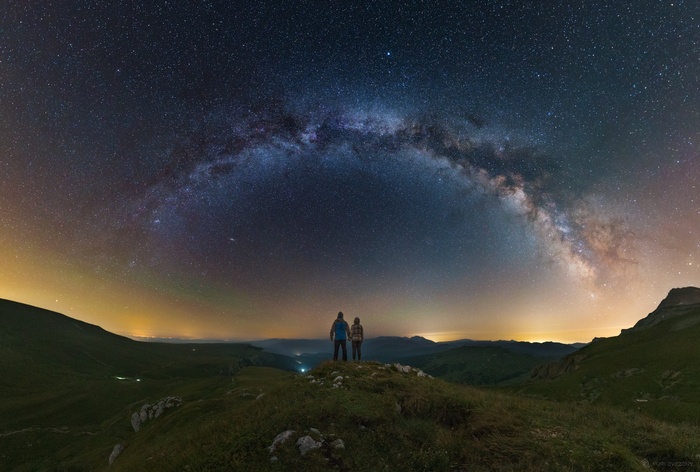 Two people stand on a hill under the Milky Way