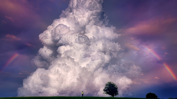 A person stands in a field with a large cloud and rainbows