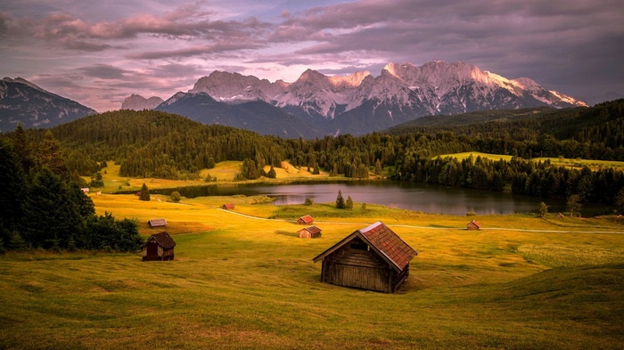 Wooden cabins in a grassy valley with mountains and a lake