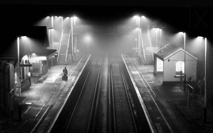 A lone figure waits at a foggy train station at night.