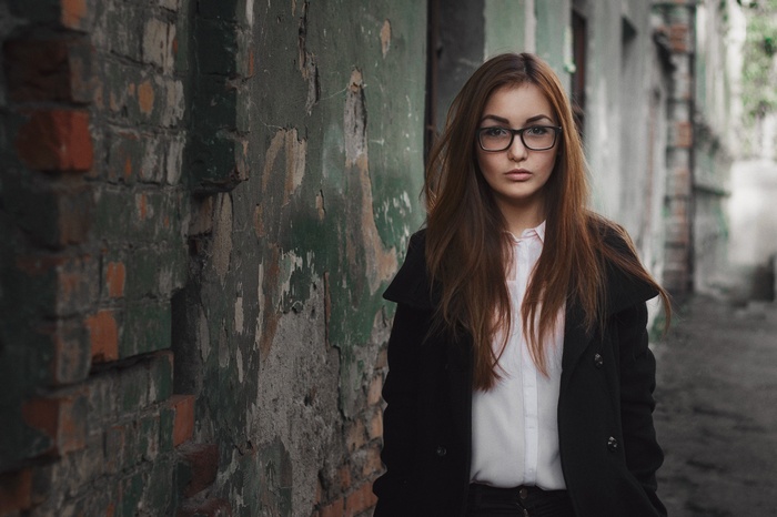 Woman with glasses and black coat standing near a brick wall