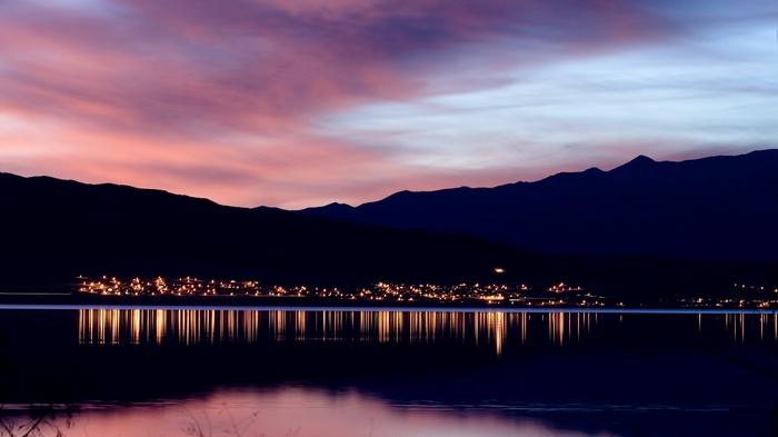 City lights reflected on a calm lake at sunset with mountains