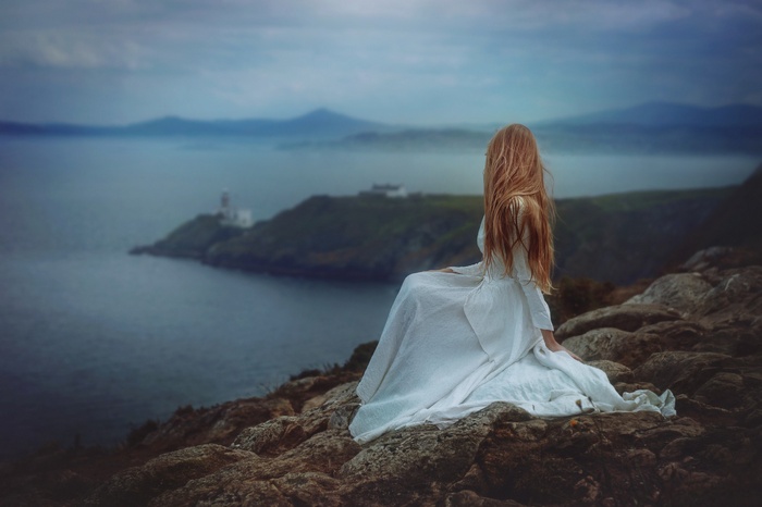 Woman in white dress on cliff overlooking sea with lighthouse