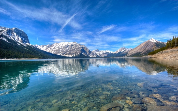 Clear lake with rocky bottom and snow-capped mountains under blue sky