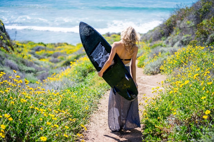 Woman with surfboard walking on a path with yellow flowers