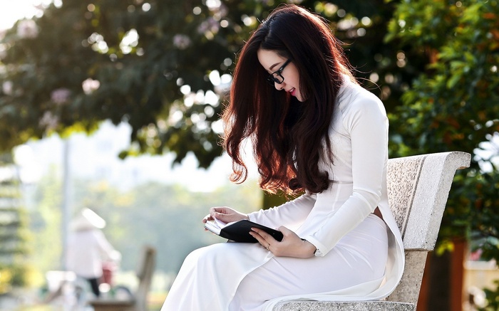 Asian woman in white dress reading a book on a bench