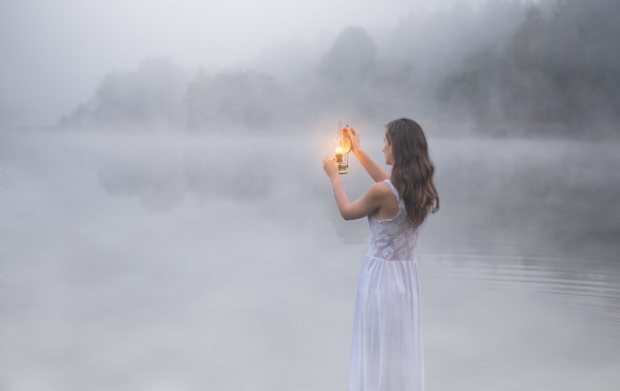 Woman in white dress holding a lantern by a misty lake