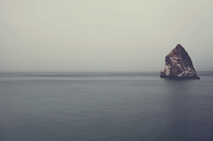 A solitary rock formation in a calm sea under a hazy sky