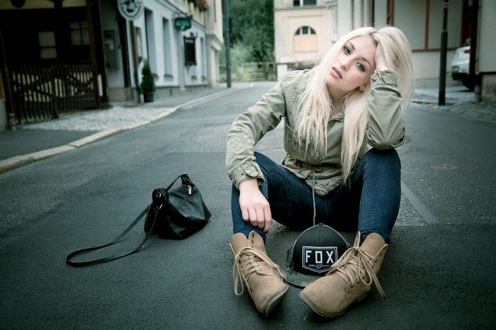 Woman in jeans and jacket sitting on road with bag and hat