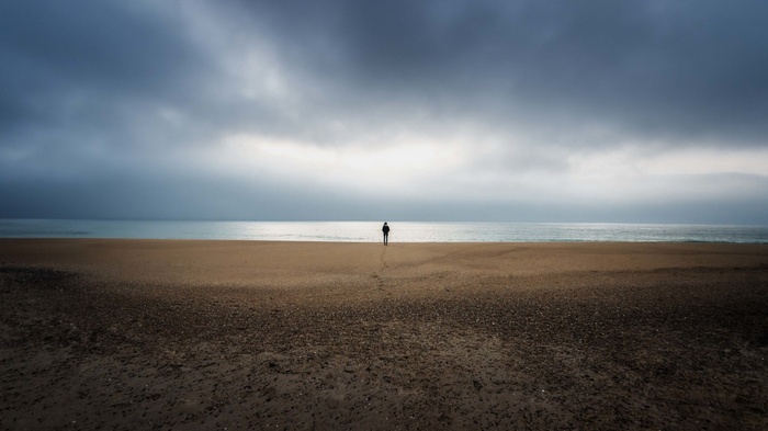 A lone figure stands on a beach facing the sea under cloudy skies.
