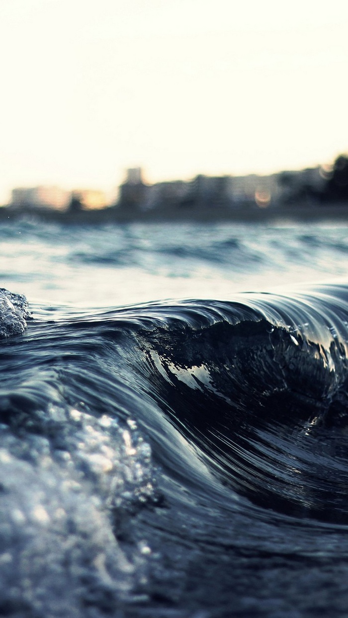 Close-up of a dark ocean wave with a blurred city skyline in the background