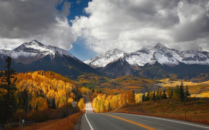Road through autumn forest to snow-capped mountains