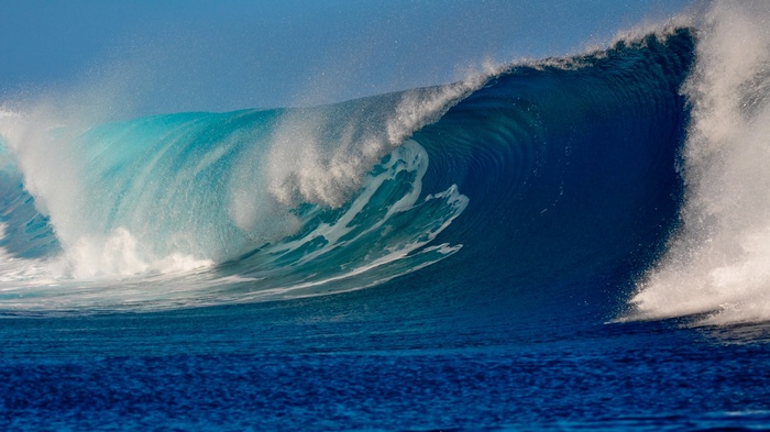 A large ocean wave breaks under a clear blue sky.