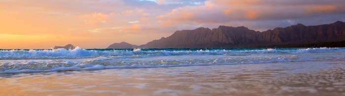 Ocean waves crash on a beach with mountains in the background at sunset.