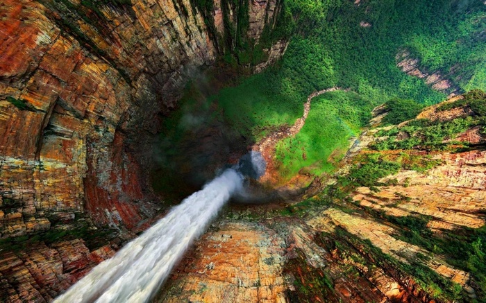 A powerful waterfall cascades into a deep canyon surrounded by lush green forest.