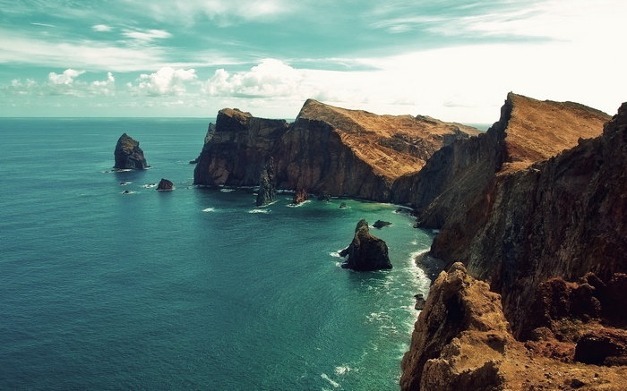Rocky cliffs meet the turquoise ocean under a cloudy sky.