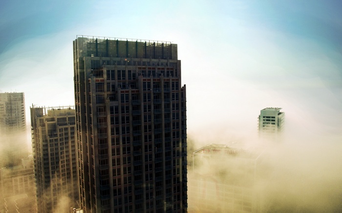Skyscrapers emerge from thick fog under a bright sky.