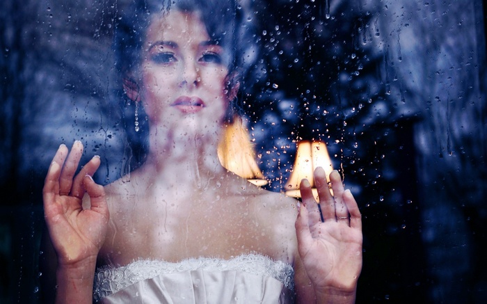 Woman looking through a rain-streaked window at a lamp