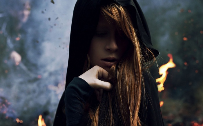 Woman with long red hair in a black hood with fire in the background