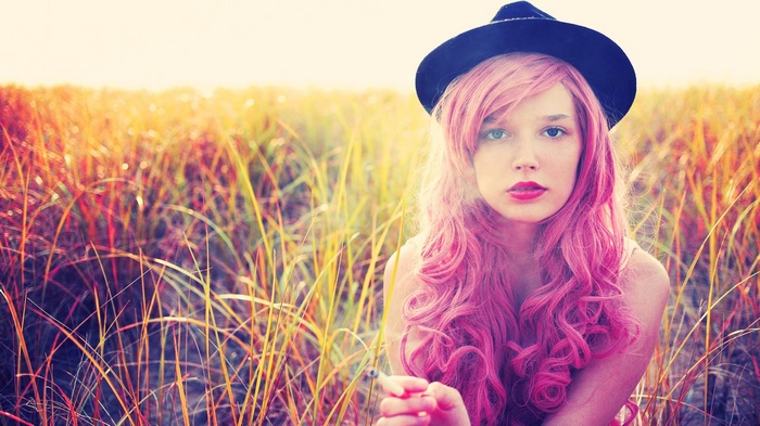 Woman with pink hair and black hat smoking in a field
