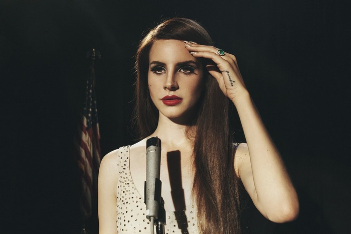 Lana Del Rey sings into a microphone with an American flag behind her