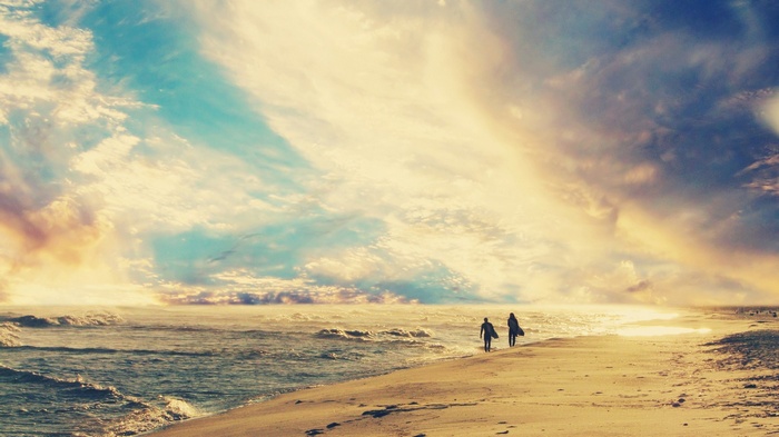 Two surfers walk along a beach at sunset