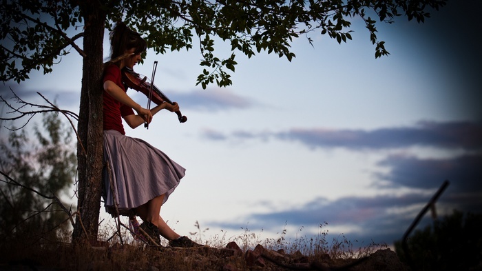 Lindsey Stirling plays violin leaning against a tree